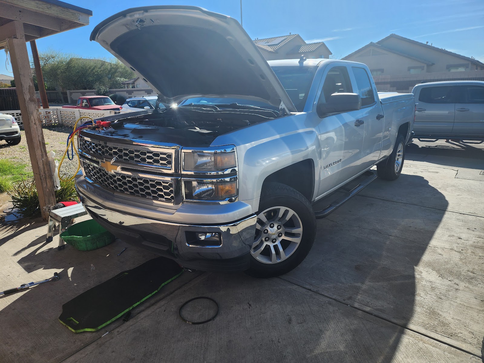 A/C gauges hooked up to a silver Silverado with the hood up, in a driveway under a clear blue sky